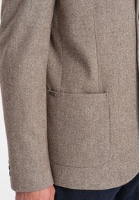 Brauner Blazer mit Fischgrätenmuster, mit aufschlag und einer Brusttasche sowie vier schwarzen Knöpfen an den Manschetten. Texturiertes Material und taillierte Passform.