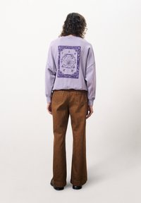 Lila Sweatshirt mit floralem grafischen Druck in Lila auf dem Rücken, kombiniert mit braunen Schlaghosen. Das Sweatshirt hat eine lockere Passform und gerippte Bündchen.