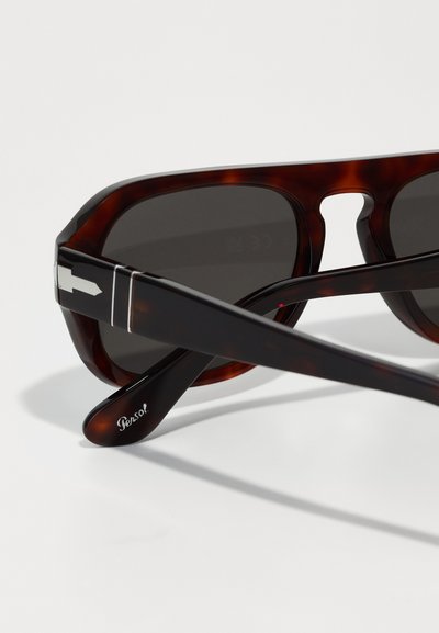 Óculos de sol em padrão tartaruga com lentes escuras, apresentando uma ponte em forma de olho de fechadura distinta, detalhes em prateado e um logótipo característico na armação.