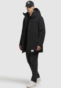 Sort parka med hætte, lynlåslukning foran, to sidelommer og et logo patch nær kanten. Sammen med mørke jeans og hvide sneakers.