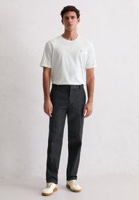 Biała koszulka z krótkim rękawem z subtelnym logo, w połączeniu z ciemnoszarymi eleganckimi spodniami i białymi tenisówkami na gumowej podeszwie, na tle jednolitego tła.