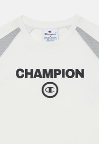 Balta Champion T-kreklu ar melniem burtiem, apaļu logotipu un gaiši pelēkām acs akcentiem uz pleciem. Klasisks apaļais kakls.