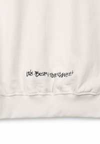 Tejido blanco con texto bordado en estilo graffiti negro que dice "NOS DIFUMINAMOS TAN DULCE" cerca del dobladillo acanalado de una prenda.