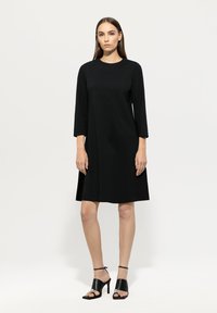 Schwarzes knielanges Kleid mit rundem Ausschnitt und Dreiviertelärmeln, aus glattem Material. Kombiniert mit schwarzen High-Heels.