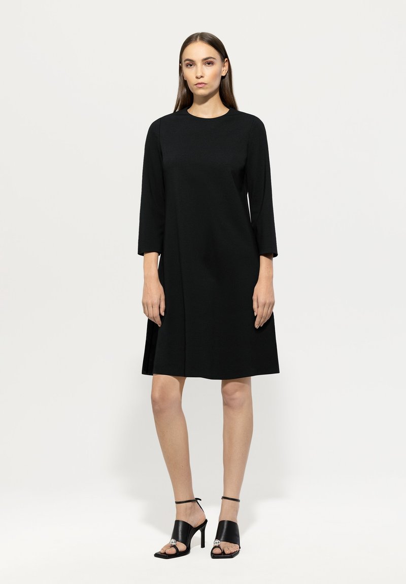 Schwarzes knielanges Kleid mit rundem Ausschnitt und Dreiviertelärmeln, aus glattem Material. Kombiniert mit schwarzen High-Heels.