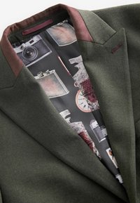 Dark green blazer with a herringbone texture, featuring a burgundy collar. Interior lining displays vintage camera and object prints.