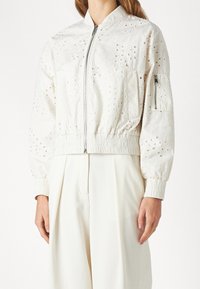 Vit bomberjacka med broderade blommönster och utskärningar, med dragkedja fram och elastiska ärmslut, bärs över vita byxor.