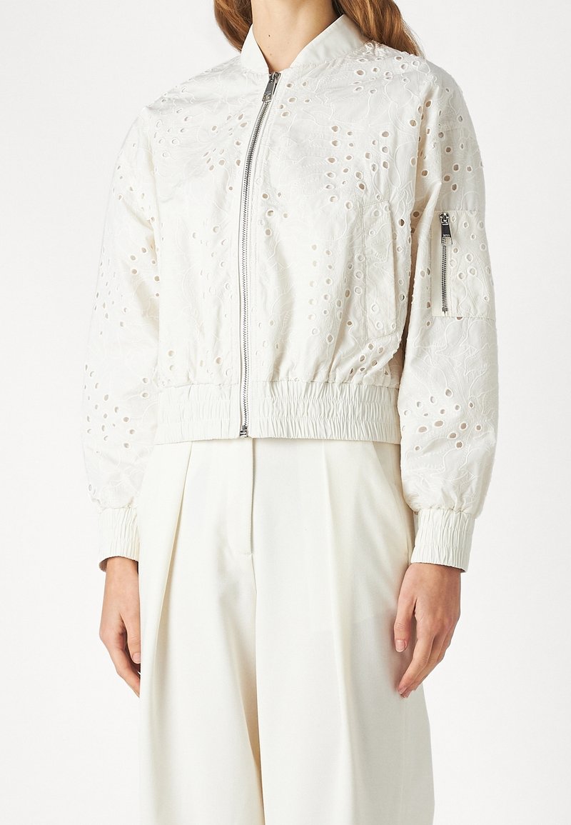 Vit bomberjacka med broderade blommönster och utskärningar, med dragkedja fram och elastiska ärmslut, bärs över vita byxor.