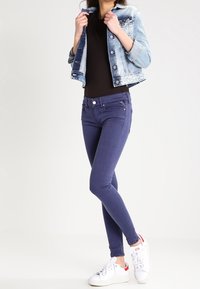 Slim-fit mörkblå jeans med framfickor och dragkedjedetalj, parat med en ljus denimjacka och vita sneakers. Svart topp under.