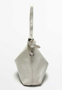 Borsa in pelle beige chiaro con una forma pentagonale unica, dotata di una singola tracolla regolabile e un accento in metallo rotondo sulla parte superiore.