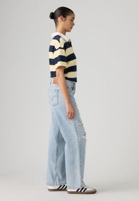 Πόλο μπλούζα με μπλε ναυτικό και κίτρινες ρίγες, με λευκό γιακά, σε συνδυασμό με ανοιχτό μπλε τζιν παντελόνι με φαρδιά μπατζάκια και σκισίματα, καθώς και μαύρα και λευκά αθλητικά παπούτσια.