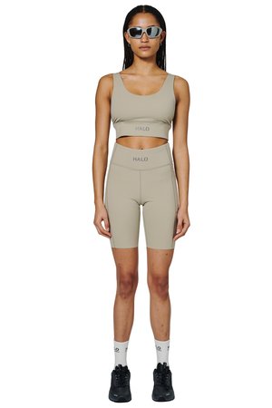 Frau trägt beigen Sport-BH und passende hoch taillierte Biker-Shorts mit "HALO"-Logo, weiße Socken, schwarze Sneakers und silberne Sonnenbrille.