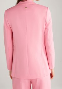 Nainen, joka on pukeutunut muotoiltuun vaaleanpunaiseen bleiseriin ja siihen sopiviin housuihin, näkyy takaa yksinkertaista vaaleaa taustaa vasten.