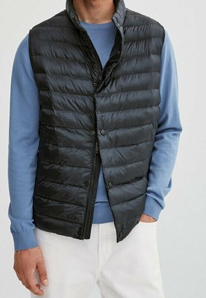 Homme portant un gilet sans manches matelassé noir par-dessus une chemise bleue à manches longues avec un pantalon blanc, main dans la poche, faisant face.