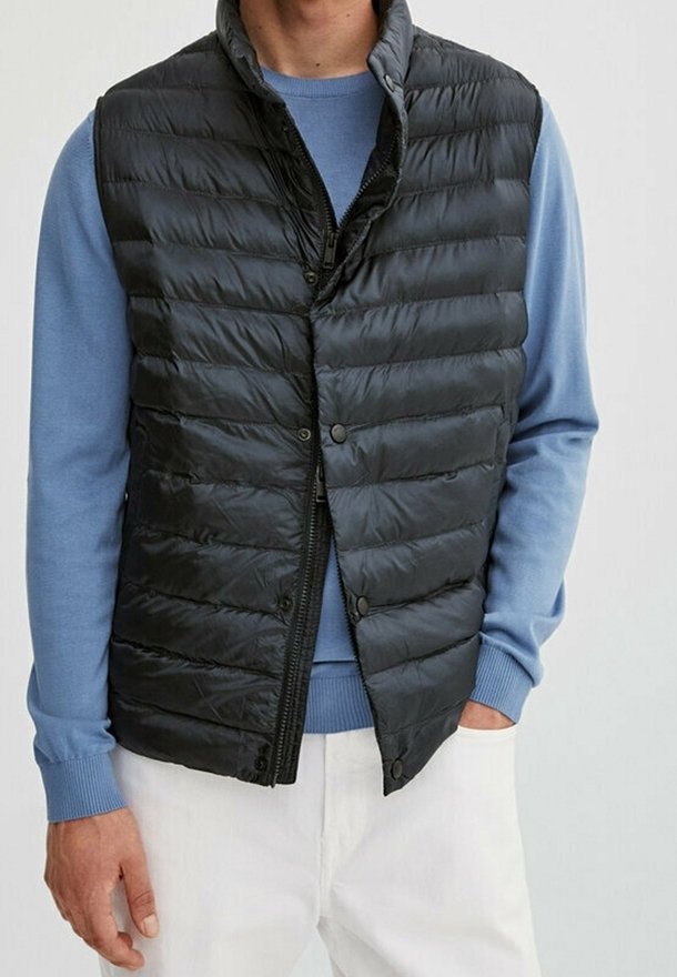 Homme portant un gilet sans manches matelassé noir par-dessus une chemise bleue à manches longues avec un pantalon blanc, main dans la poche, faisant face.