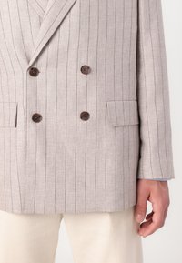 Double-breasted linen blazer in light beige with dark pinstripes. Features brown buttons, notched lapels, and side pockets. Textured fabric.