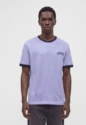 Jeune homme portant un T-shirt violet clair avec bordure noire et le mot "DOUBLE A" imprimé sur la poitrine, debout devant un fond uni.