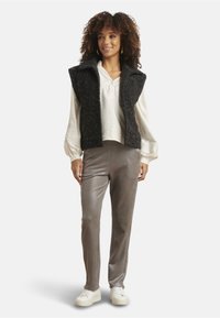 Schwarze, strukturierte Weste über einer weißen Bluse, kombiniert mit glänzenden grauen Hosen und weißen Turnschuhen. Die Weste hat einen Kragen und eine Knopfleiste.