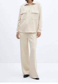 Jersey de punto beige con cremallera en el cuello, dos bolsillos en el pecho y mangas largas, combinado con pantalones beige de pierna ancha, con una textura suave.