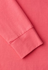 Korallfärgad långärmad skjorta med ett mjukt, texturerat tyg och en uppvikt manschett. Designen är enkel, utan synliga mönster eller dekorationer.