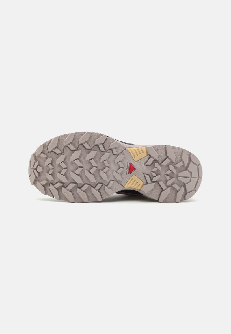 Suela de zapato de senderismo con profundos surcos angulares en gris claro y negro. Presenta un acento triangular en rojo y componentes de goma amarilla.