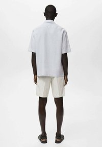 Lys grå kortærmet skjorte, enkel design, afslappet pasform, parret med hvide skræddersyede shorts. Båret med sorte sandaler. Baggrunden er ensfarvet.