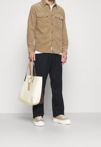 Camisa de pana marrón con dos bolsillos en el pecho, pantalones anchos azul marino, zapatillas beige y un bolso tote crema y marrón con detalles de cuero.
