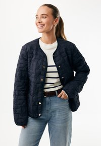 Granatowa pikowana kurtka z okrągłym kołnierzem, ozdobiona złotymi zatrzaskami i teksturowanym wzorem. Noszona na białym swetrze w granatowe paski.