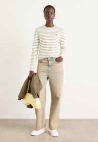 Camicia a maniche lunghe a righe in crema e oliva, abbinata a jeans a gamba larga in denim chiaro. Accessoriata con una borsa gialla e un giubbotto marrone.