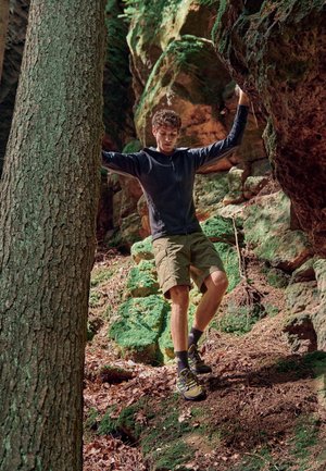 Outdoor-Abenteuer-Outfit mit einer schwarzen Reißverschlussjacke, grünen Shorts und gelben Wanderschuhen. Natürliche Waldumgebung mit Felsen und Moos.