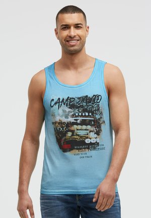 Man wearing light blue tank top with off-road vehicle graphic and text, smiling, standing against plain light background.