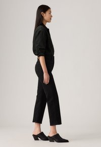 Jeans neri in denim a vita alta abbinati a una camicia verde scuro con bottoni. Modella che indossa mules nere con tacco. Vista di profilo.