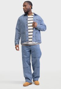 Denim jacket in light blue, button-up front, two chest pockets. Striped shirt beneath, wide-leg denim pants, and tan footwear.