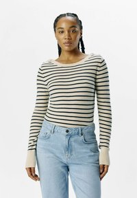 Beige og marineblå stribet striktrøje med lange ærmer og ribbet manchetter. Har en bred rund halsudskæring med knapper på skulderen. Bæres med lyse jeans.