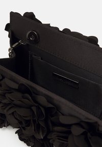 Sac à main en tissu noir avec des accents de fleurs texturées, une fermeture à pression, et une poche intérieure avec une plaque logo. Design lisse et minimaliste.