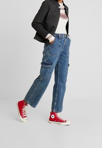 Pantalon cargo en denim bleu à coupe ample, doté de plusieurs poches latérales et de passants pour ceinture. Associé à des baskets montantes rouges et une veste noire.