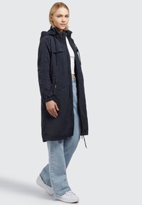 Giacca lunga blu navy con cappuccio, realizzata in materiale leggero. Presenta due tasche laterali, coulisse regolabili e zip frontale.