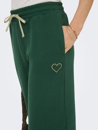 Pantalon de jogging vert avec taille élastique et cordon doré, main dans la poche, détail cœur brodé en or et deux bracelets dorés au poignet.