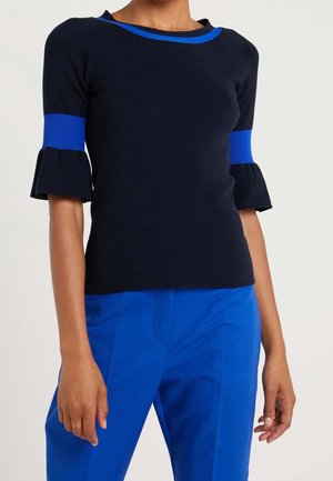 Personne portant un haut noir ajusté avec une bordure bleue au niveau de l'encolure et des manches, associé à un pantalon bleu vif, debout devant un fond uni.