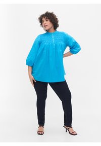 Turkizna bluzica s napuhanim rukavima i detaljima dugmadi sprijeda. Ima nabranu površinu na grudima. Uparena s tamnim trapericama i potpeticama.