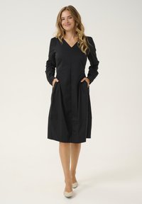 Crna haljina s dugim rukavima i V izrezom, koja ima bočne džepove i lepršavi kroj. Glatka tekstura i dužina do koljena.