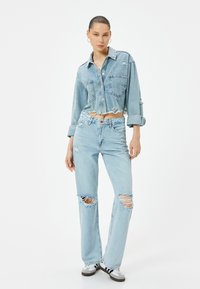 Chaqueta de mezclilla azul claro corta con cuello puntiagudo y bolsillos en el pecho, combinada con jeans de tiro alto que presentan desgastes en las rodillas.