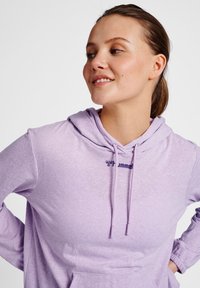 Ljuslila huvtröja i mjukt, texturerat tyg. Har en dragsko vid huvan och en liten marinblå logotyp på bröstet. Långa ärmar.