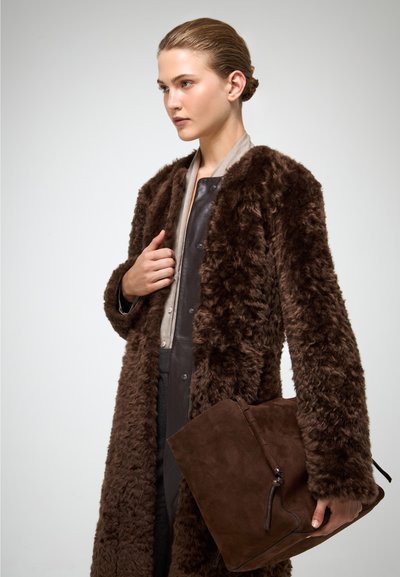 Bruine faux fur jas met een lange silhouet en een gestructureerd oppervlak. Het model houdt een donkerbruine suède tas vast met een striksluiting.