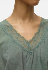 Top verde a collo a V con delicata rifinitura in pizzo attorno al collo. Il tessuto ha una consistenza morbida e una leggera lucentezza.