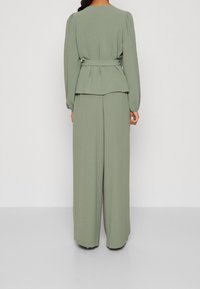 Completo a due pezzi verde e texturizzato con vita peplum e pantaloni a gamba larga. Maniche lunghe, scollatura a giro e vita regolabile accentuano la silhouette.