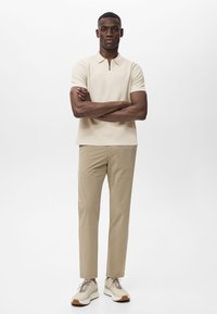 Beige poloko s krátkym rukávom a polozipsovým golierom, kombinované so svetlo khaki nohavicami a béžovými teniskami s hnedými akcentmi. Jednoduchý, moderný strih.