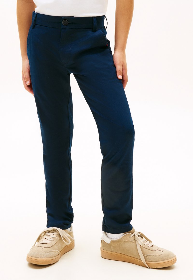 Enfant portant un pantalon bleu foncé, des baskets beiges avec des chaussettes blanches, debout les mains détendues le long du corps sur un fond uni.