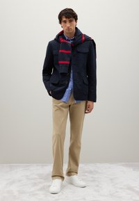 Mann mit marineblauer Jacke, blau gestreiftem Hemd, beigen Hosen und weißen Turnschuhen, mit einem navy und rot gestreiften Pullover über den Schultern.