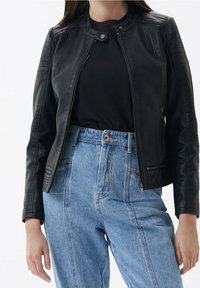 Donna che indossa una giacca di pelle nera sopra una maglietta nera con jeans a vita alta di un blu chiaro, in piedi contro uno sfondo bianco semplice.